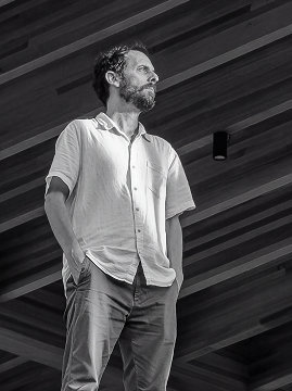 Retrato em preto e branco de um homem de barba e camisa clara, sob forro ripado de madeira.