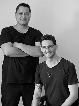 Retrato em preto e branco de dois homens de camiseta preta, um deles de braços cruzados.